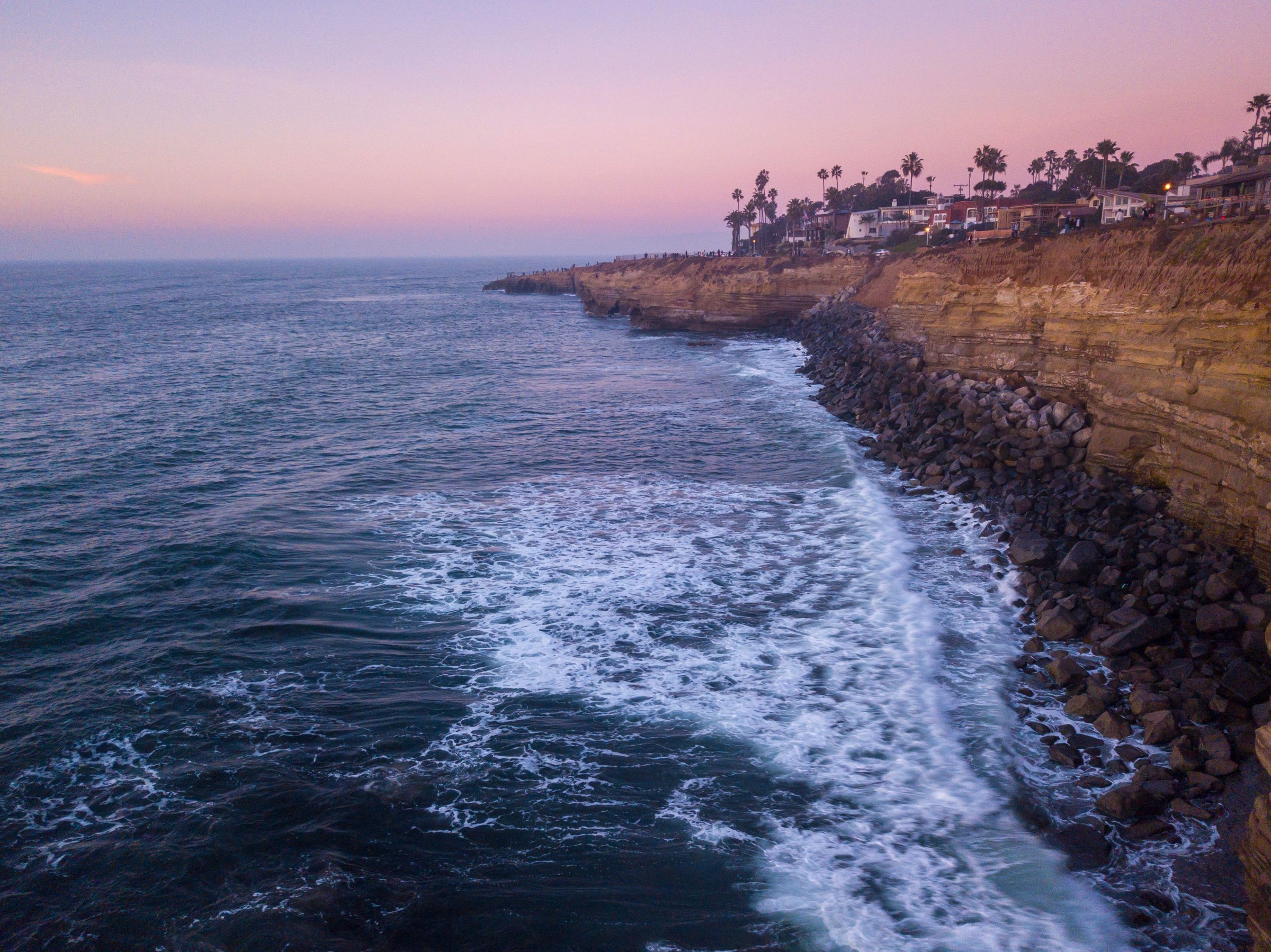Aerial view at dusk of tall tan sandstone cliffs along the ocean, with white waves breaking on dark rocks at the base and a pink and purple sky