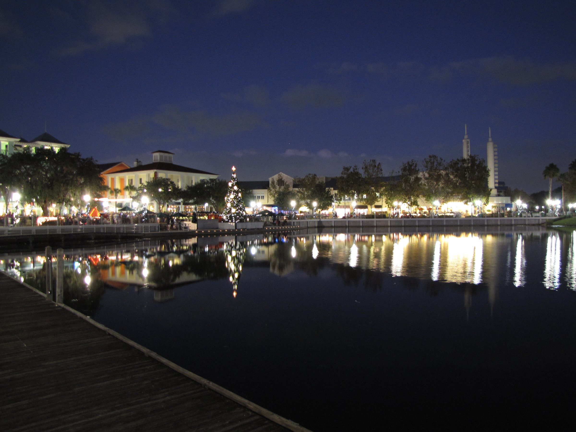 Warm lights from low buildings, a small lit Christmas tree and two white spires reflect in a dark lake at night