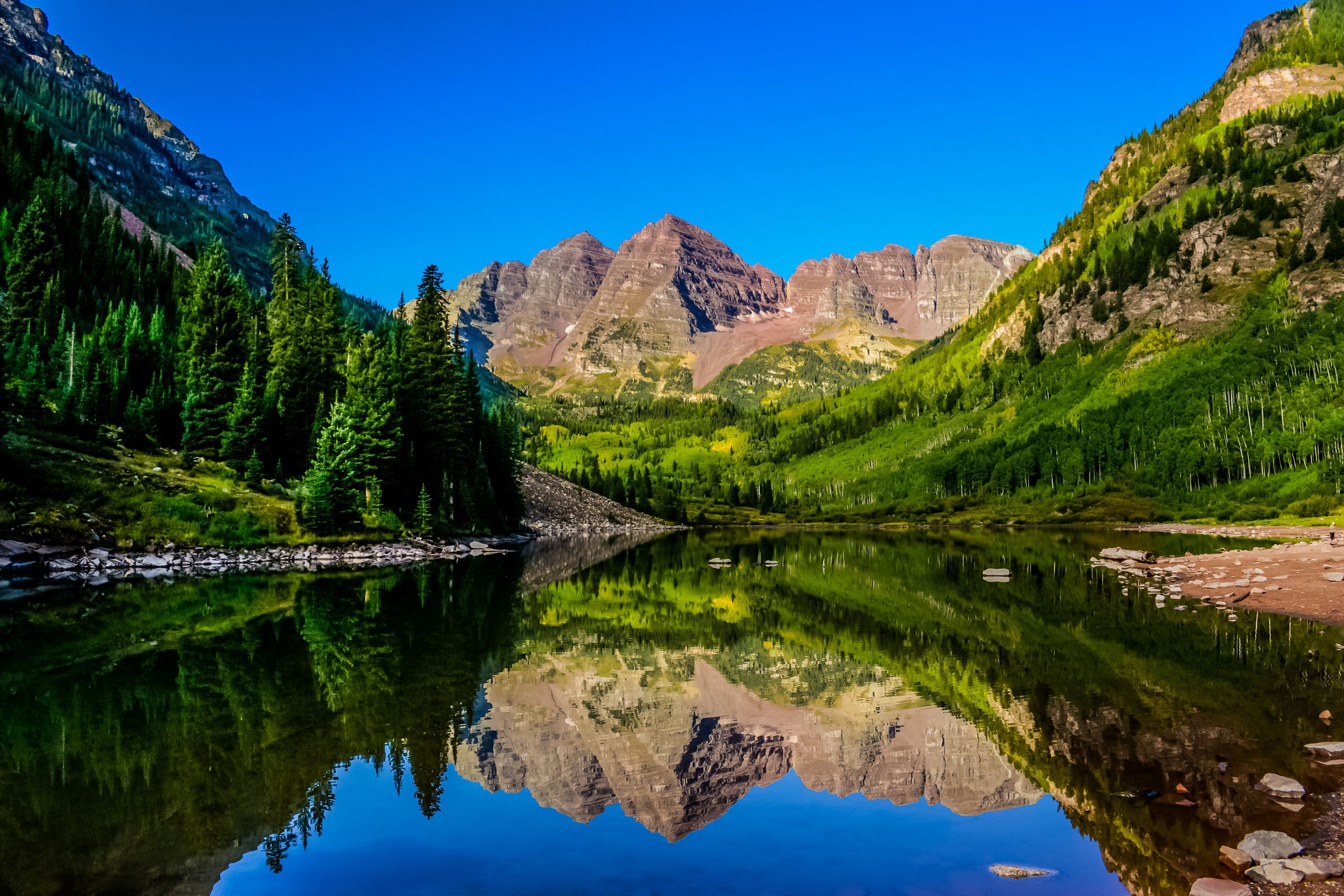 Two reddish mountain peaks at the head of a green valley, reflected in a still lake, with evergreen trees on both sides under a deep blue sky