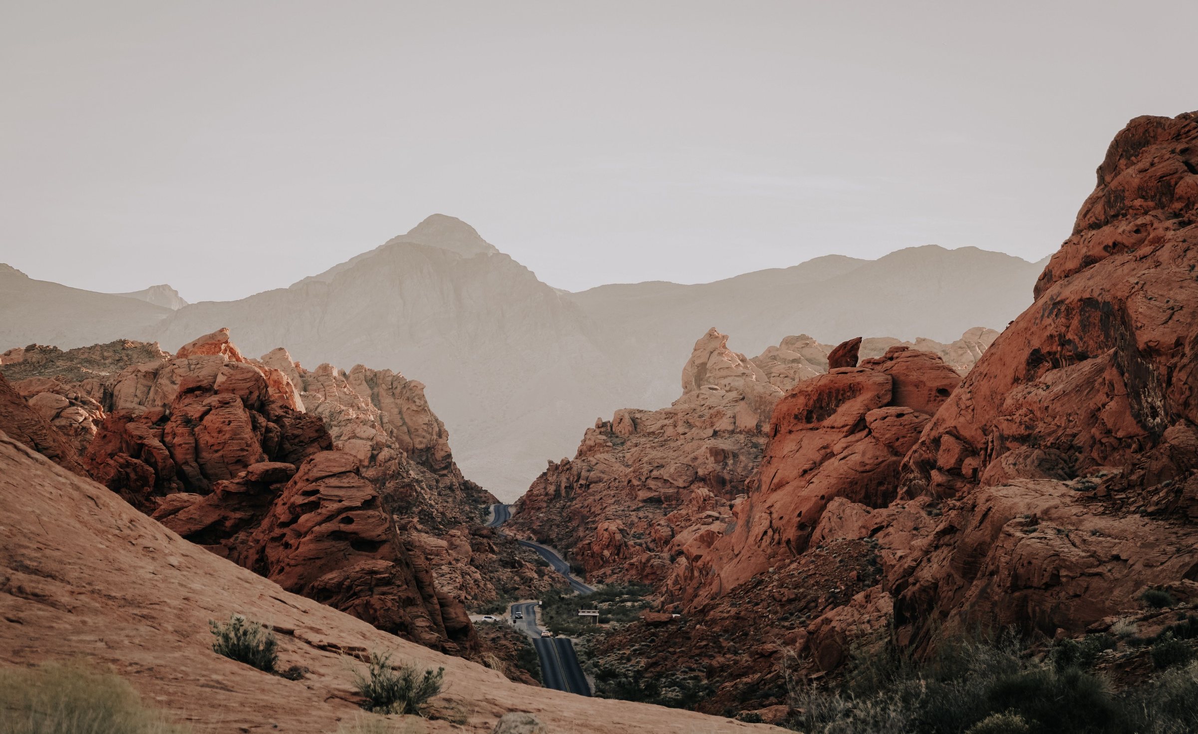 Red and orange sandstone rock formations and rocky slopes, with gray mountains behind them under a hazy sky