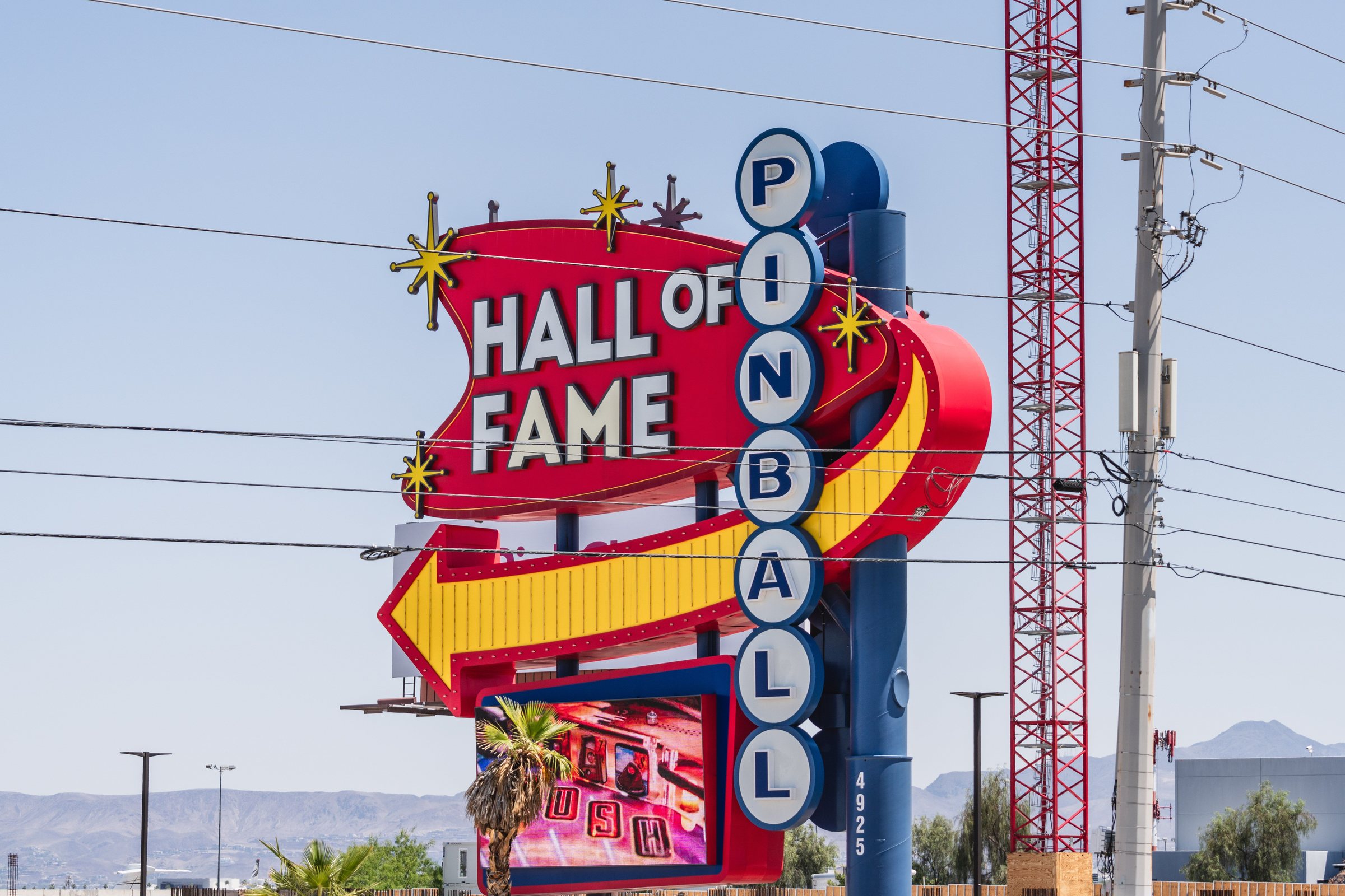 A tall sign with PINBALL in blue circles, HALL OF FAME in red and a yellow arrow, against a pale daytime sky with power lines