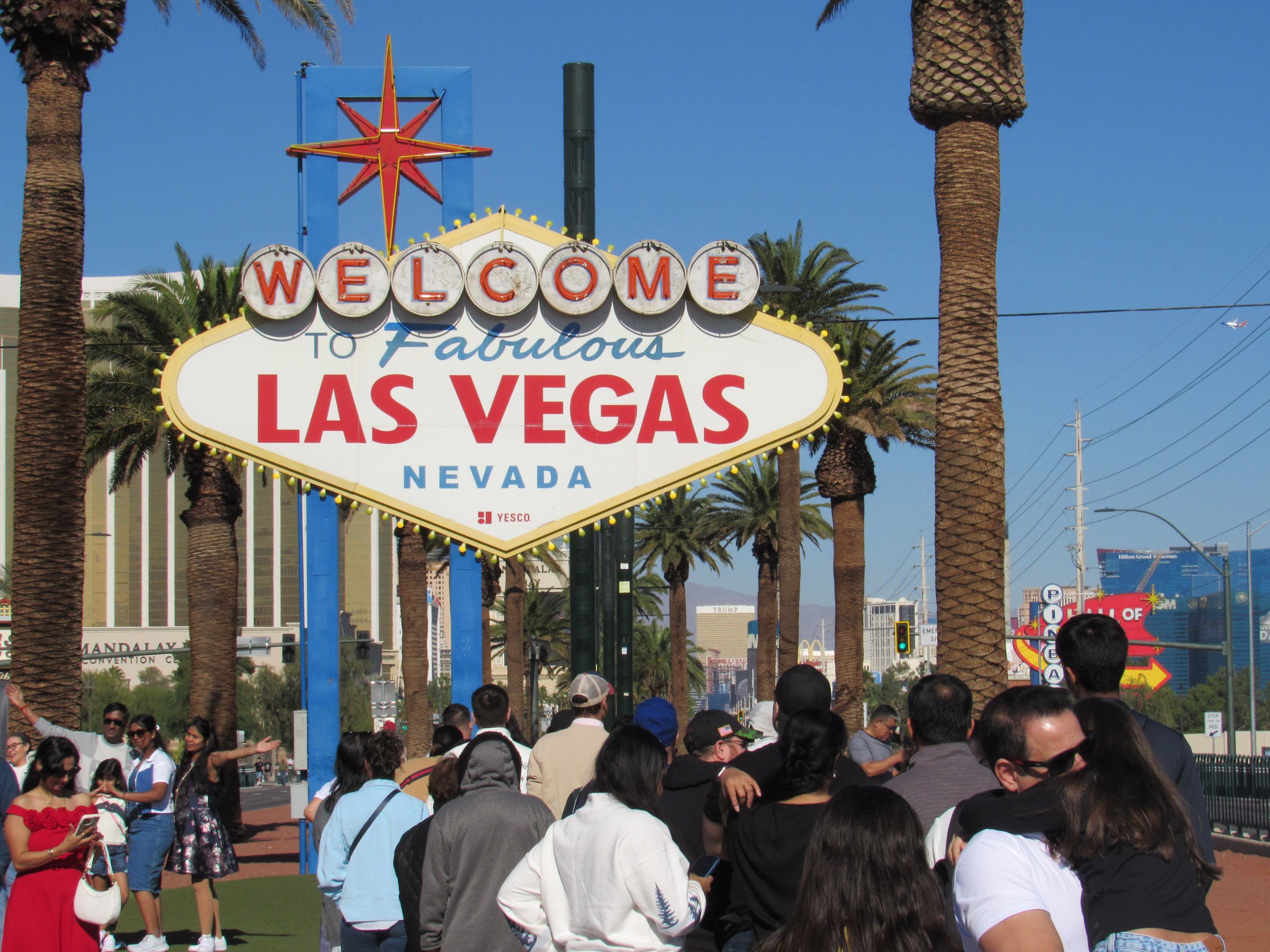 A crowd of people in front of the sign reading WELCOME to Fabulous LAS VEGAS NEVADA, with palm trees around it and a blue sky