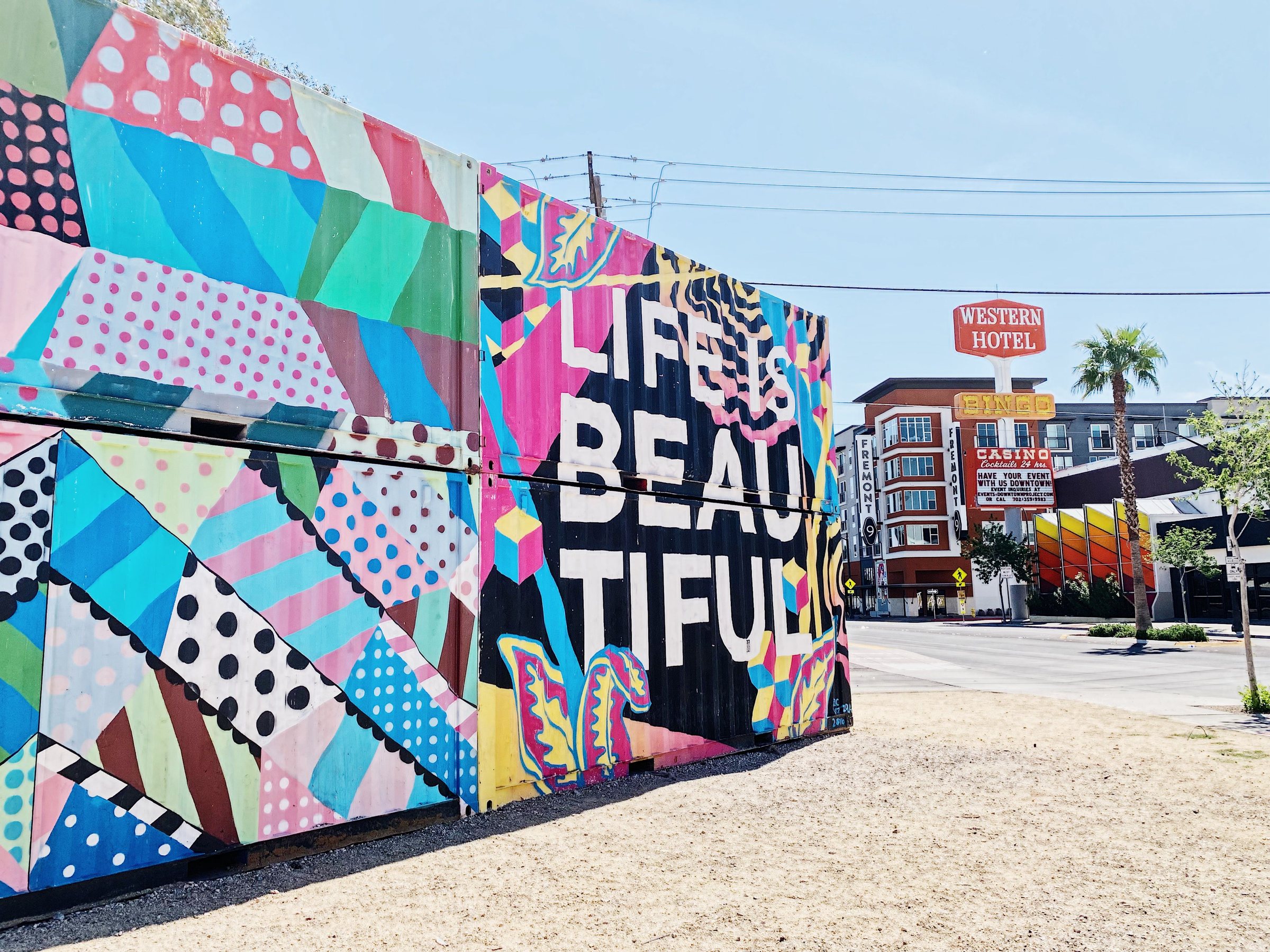 A wall covered in a brightly colored patchwork mural with the words LIFE IS BEAUTIFUL, with a palm tree and a small red sign reading Western in the distance