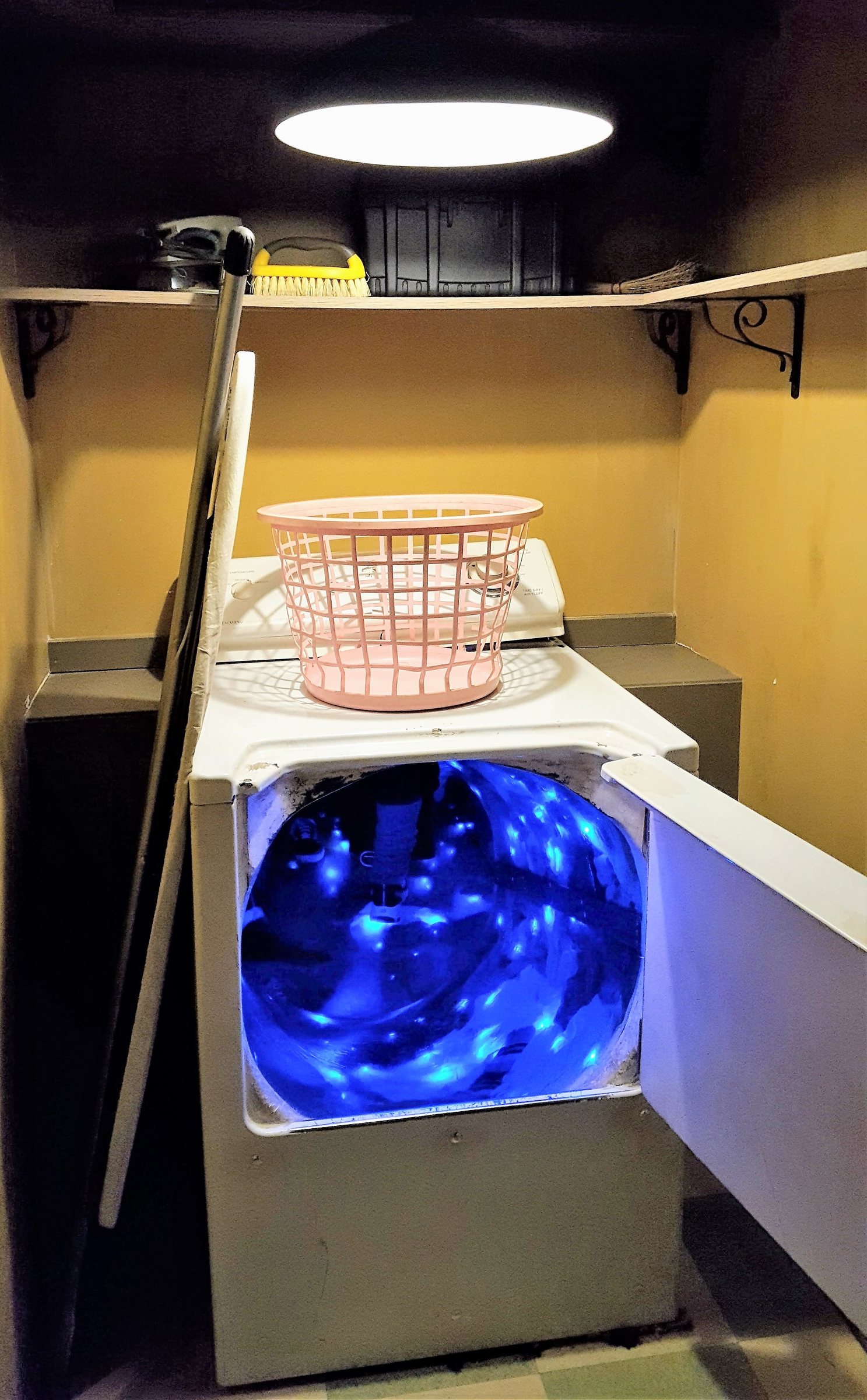 An open white washing machine glowing blue inside, with a laundry basket on a shelf above it