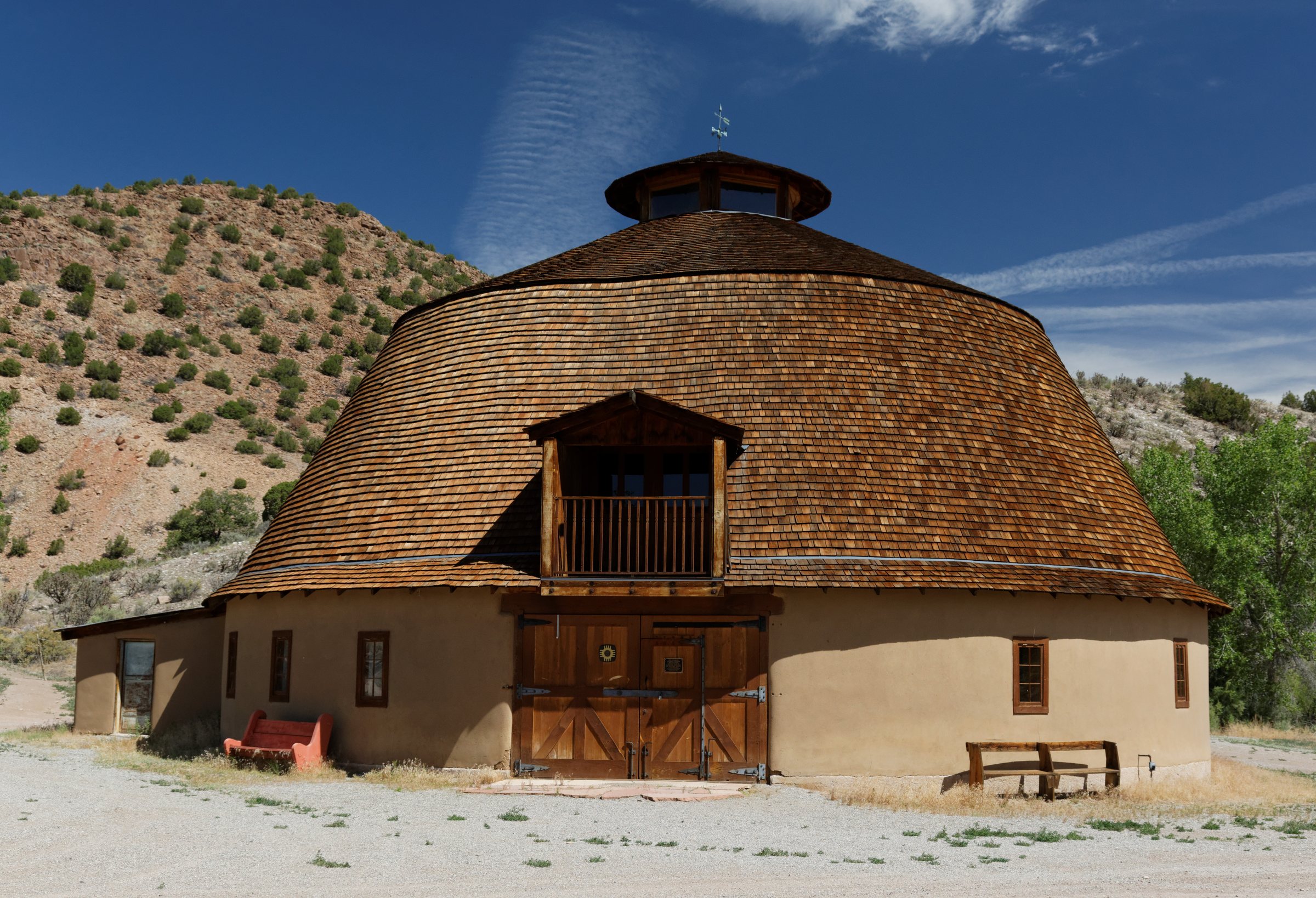 A large round barn with a wooden shingle roof and a cupola under a blue sky with clouds, at Ojo Caliente
