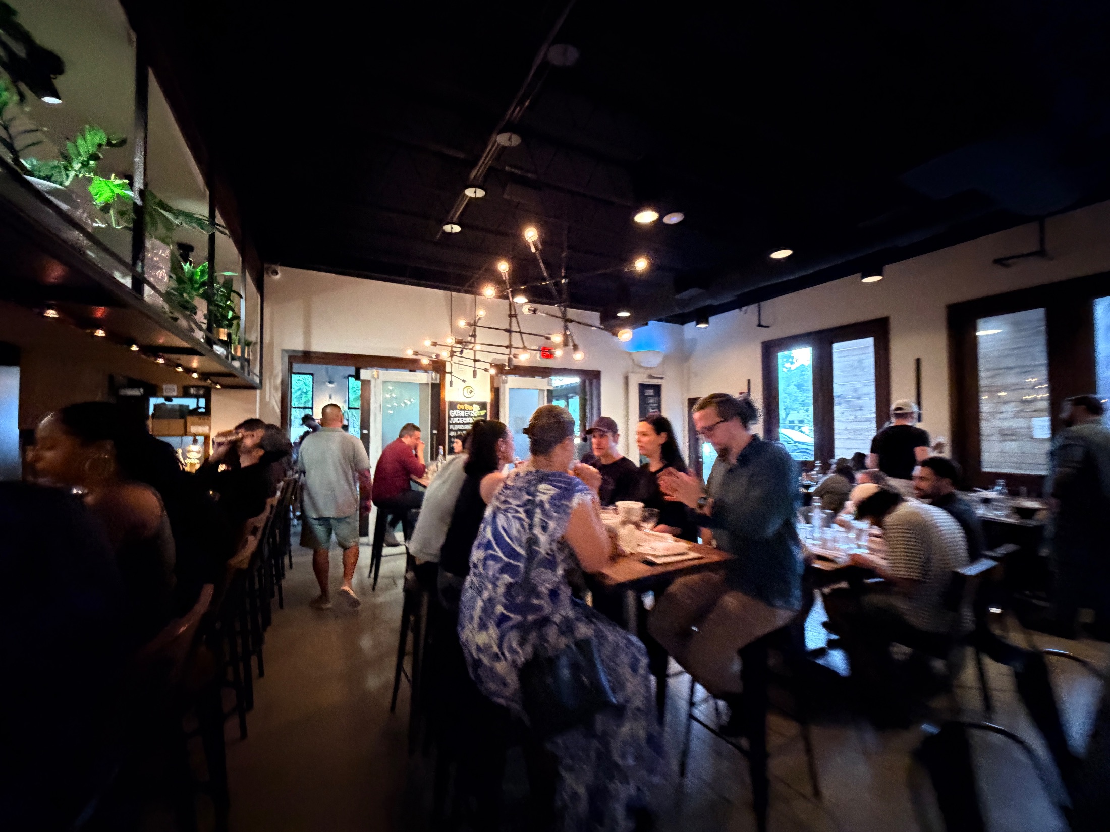 View through DOMU's entrance into the dining room, showing communal tables, hanging plants on an iron rail, a ramen mural on the far wall, and the Buy the Kitchen a Round sign on the right