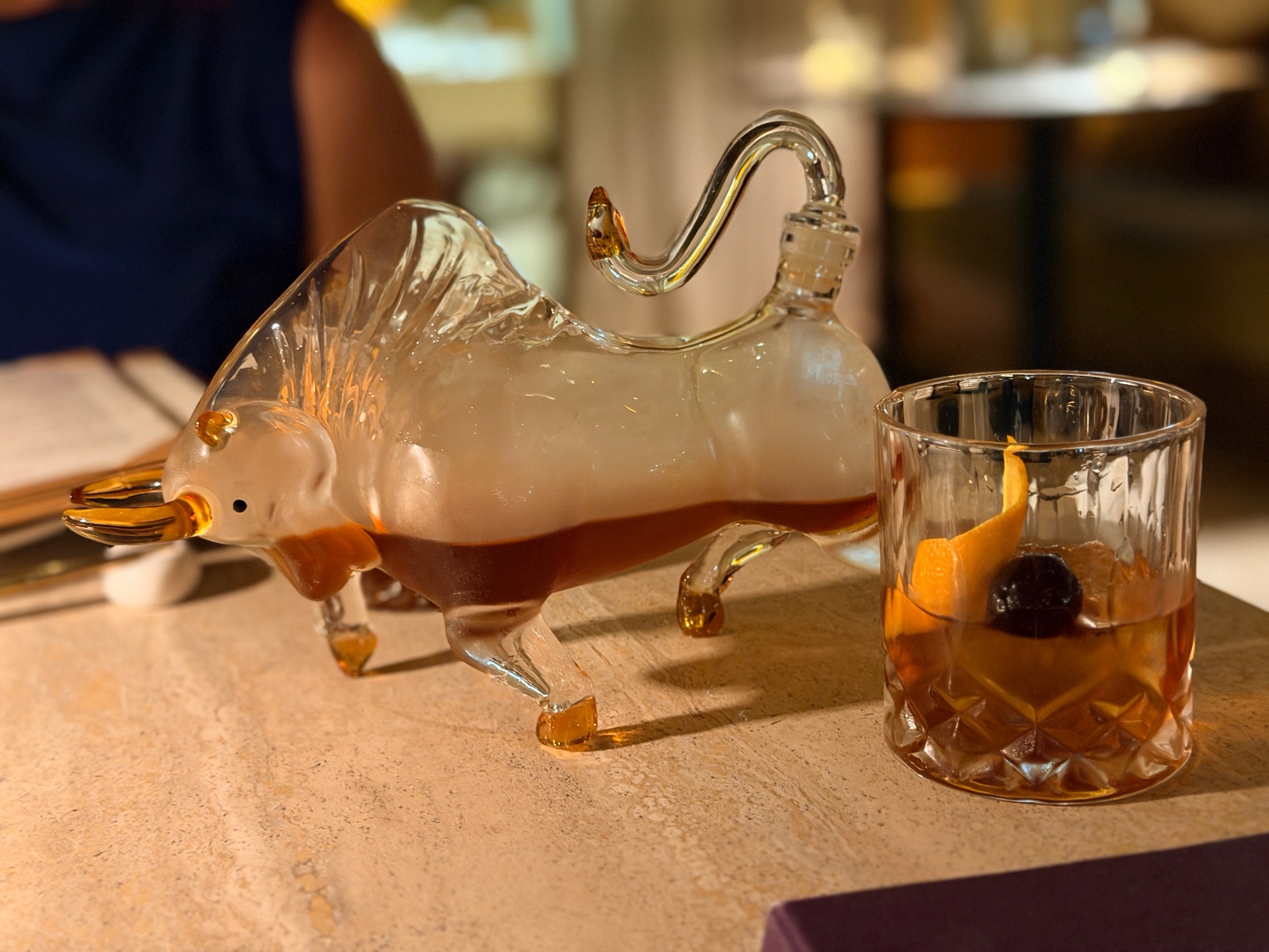 The Taurus in Retrograde cocktail served in a glass bull decanter alongside a rocks glass with orange peel