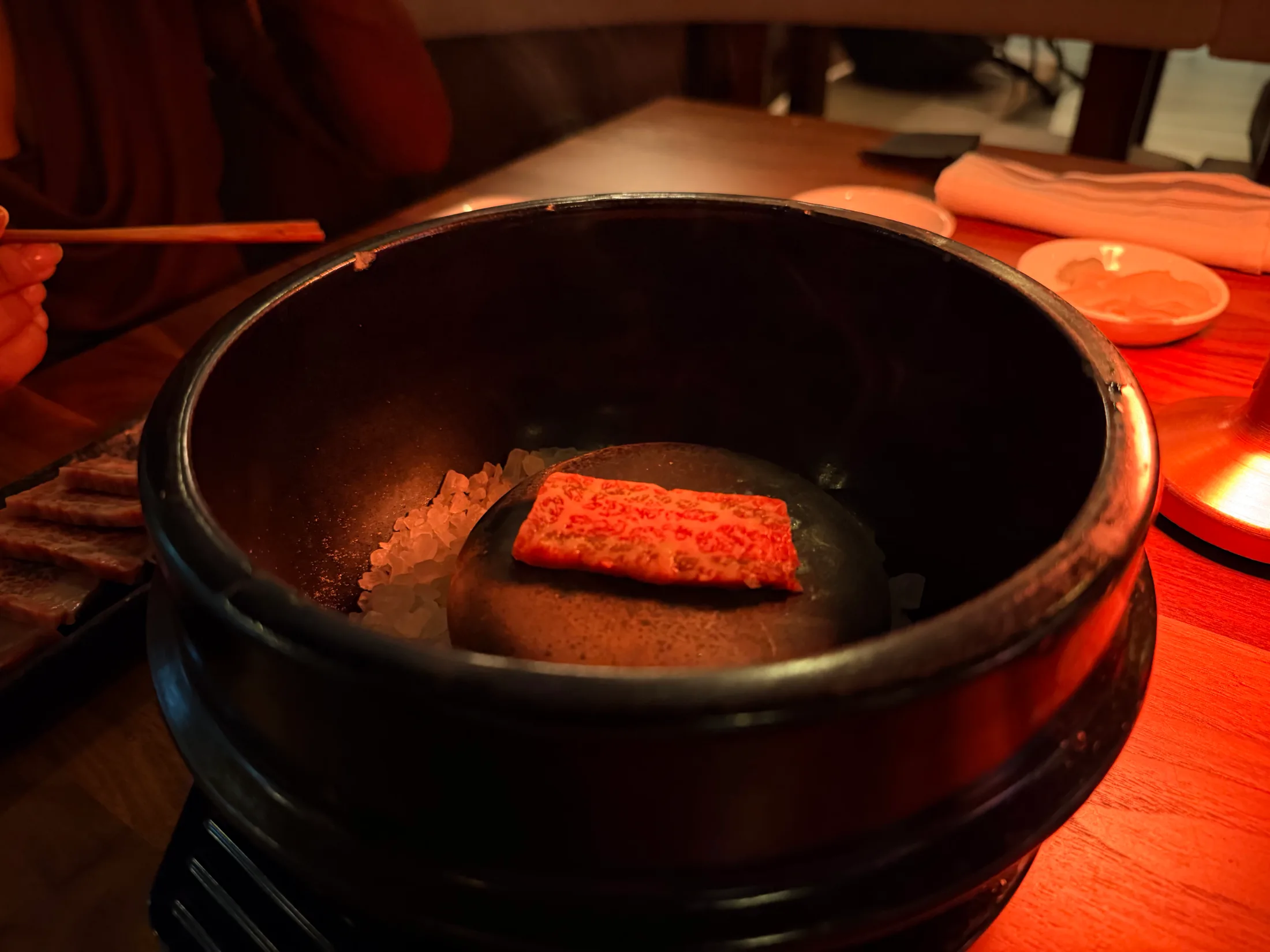 A single vivid piece of nigiri resting on rock salt inside a dark ceramic vessel, lit by warm red ambient light