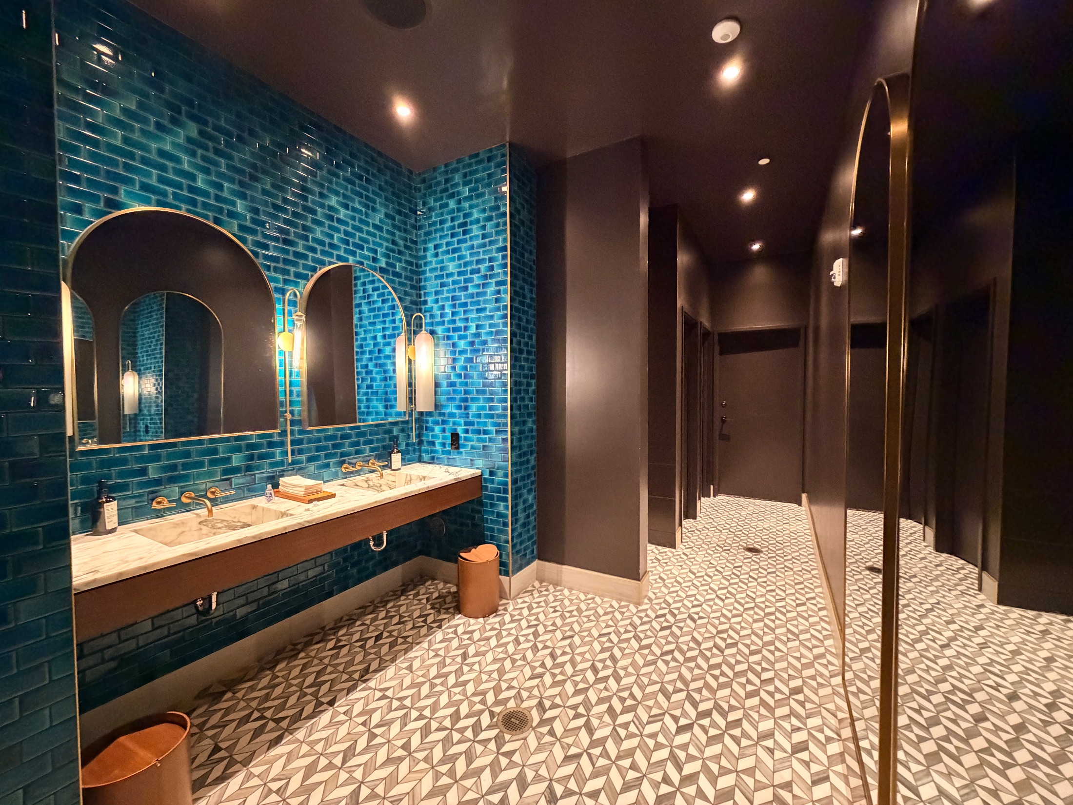 Deep teal subway tile bathroom with brass arched mirrors and geometric patterned floor at Salt and the Cellar
