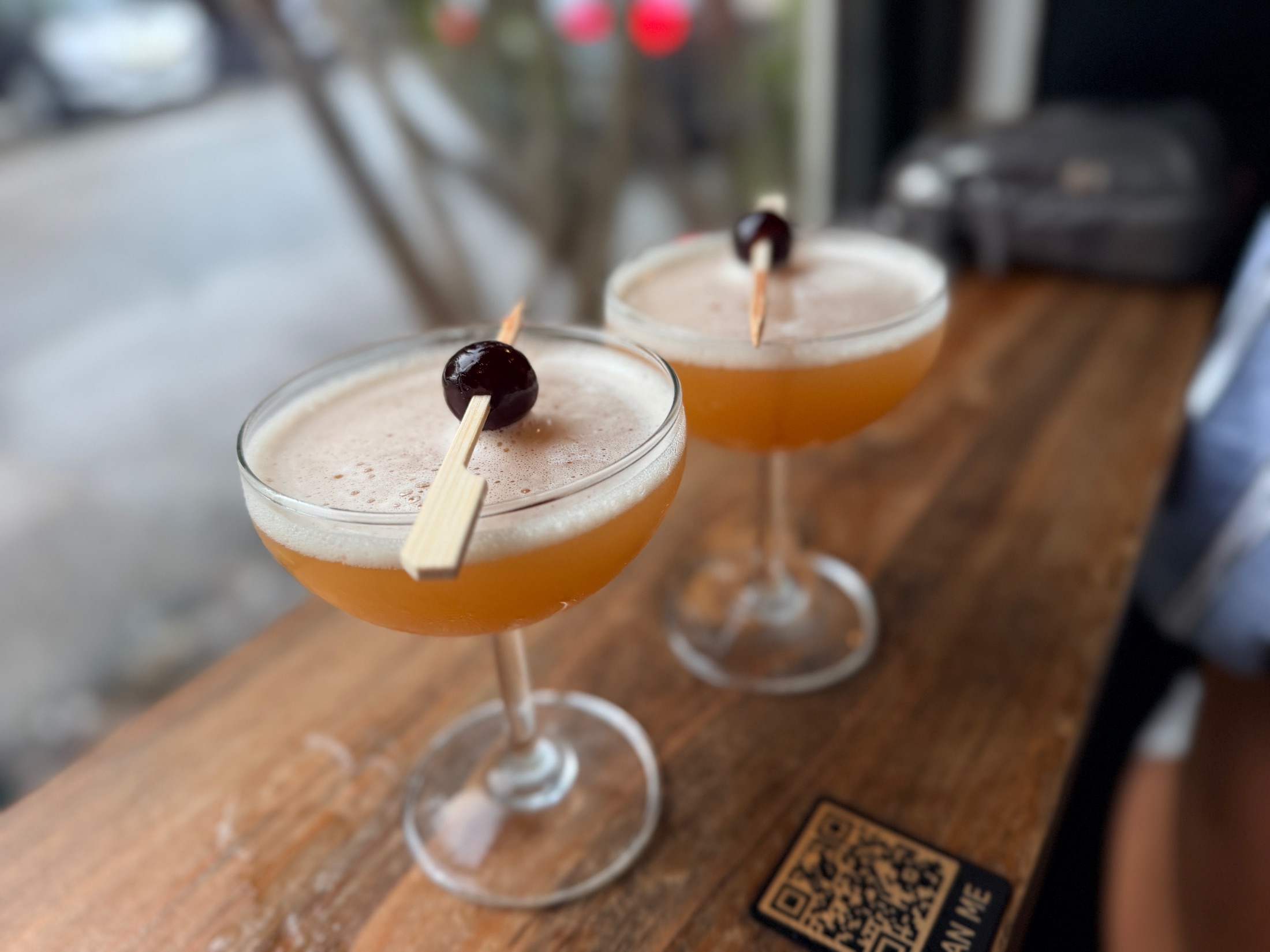 Two amber-colored cocktails in coupe glasses, each with a dark cherry on a toothpick, resting on a wood surface at DOMU