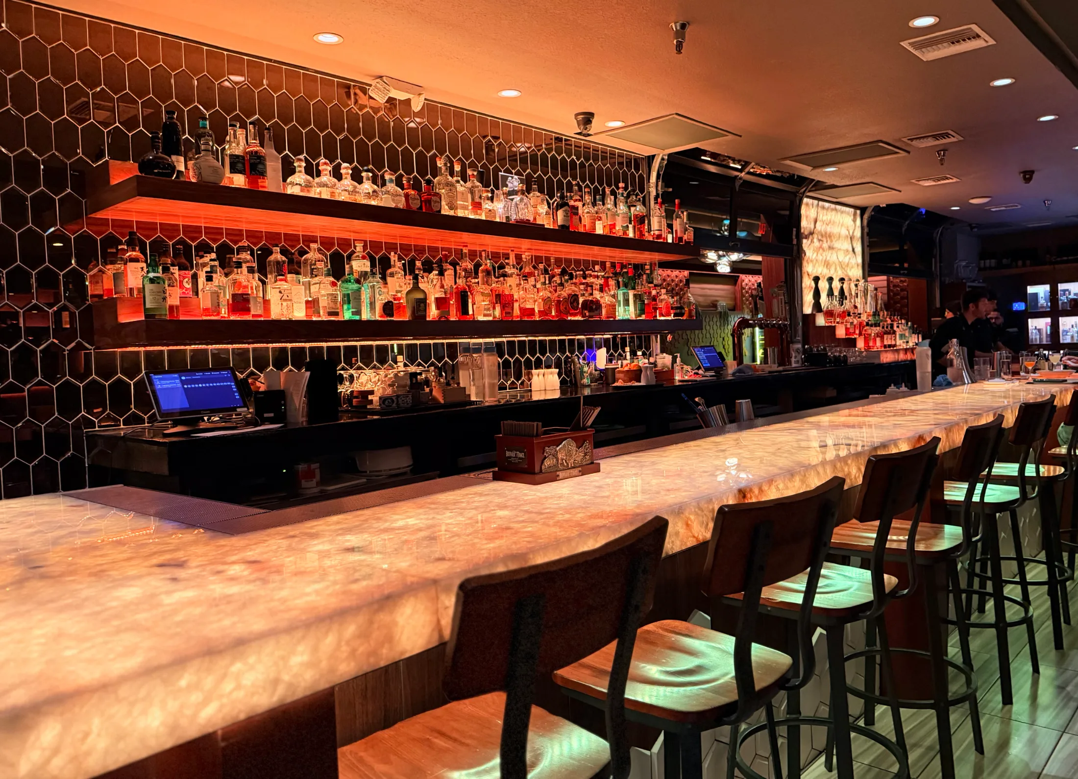 Long marble bar counter with warm amber lighting, honeycomb tile backsplash, and shelves lined with premium spirits bottles
