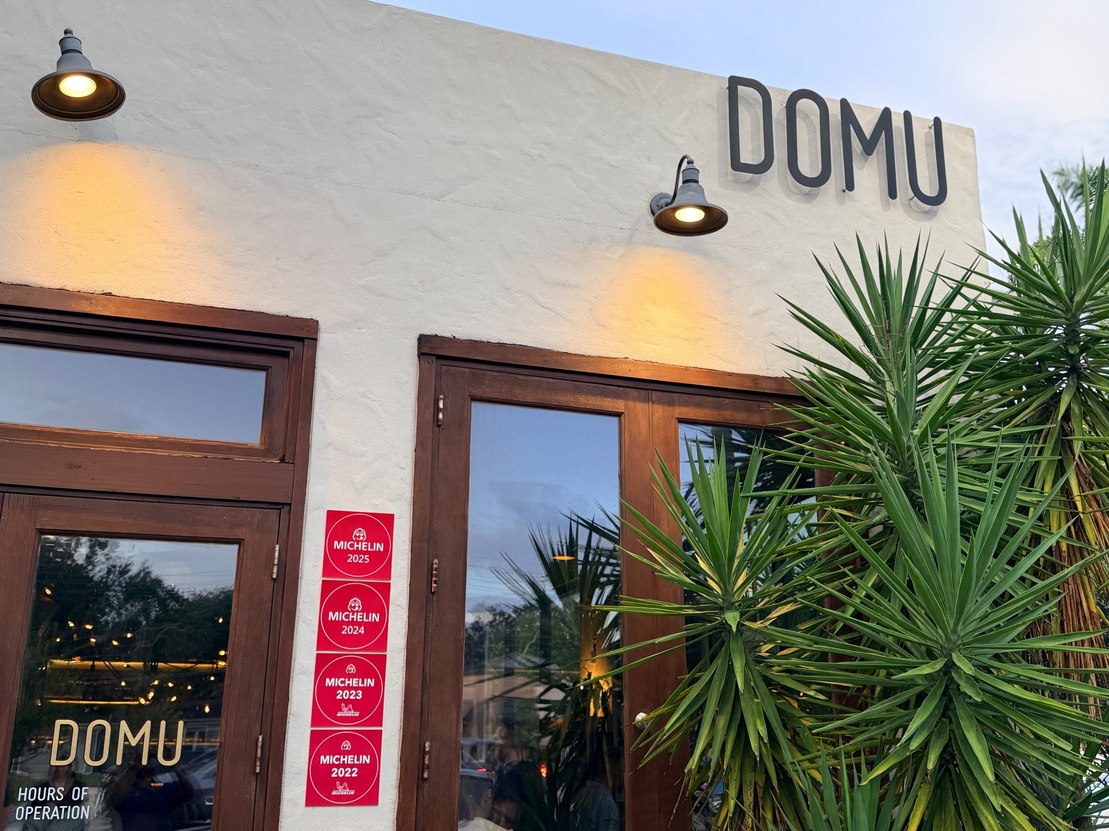 DOMU's white stucco exterior at dusk with four red Michelin Bib Gourmand plaques stacked beside the door, 2022 through 2025