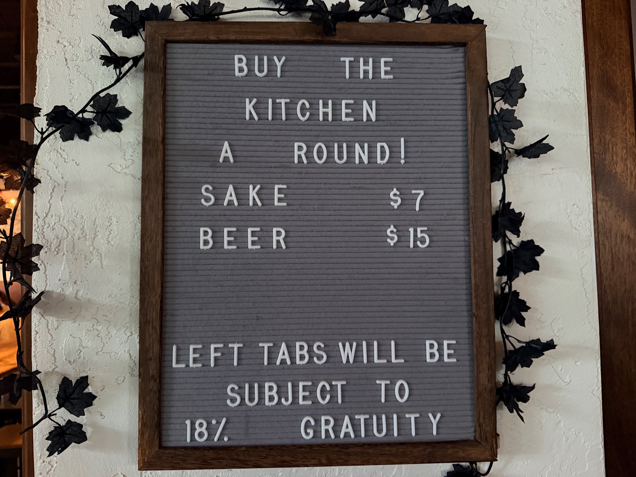 A gray letter board sign reading BUY THE KITCHEN A ROUND, SAKE $7, BEER $15, LEFT TABS WILL BE SUBJECT TO 18% GRATUITY, framed in wood and surrounded by faux vine decor