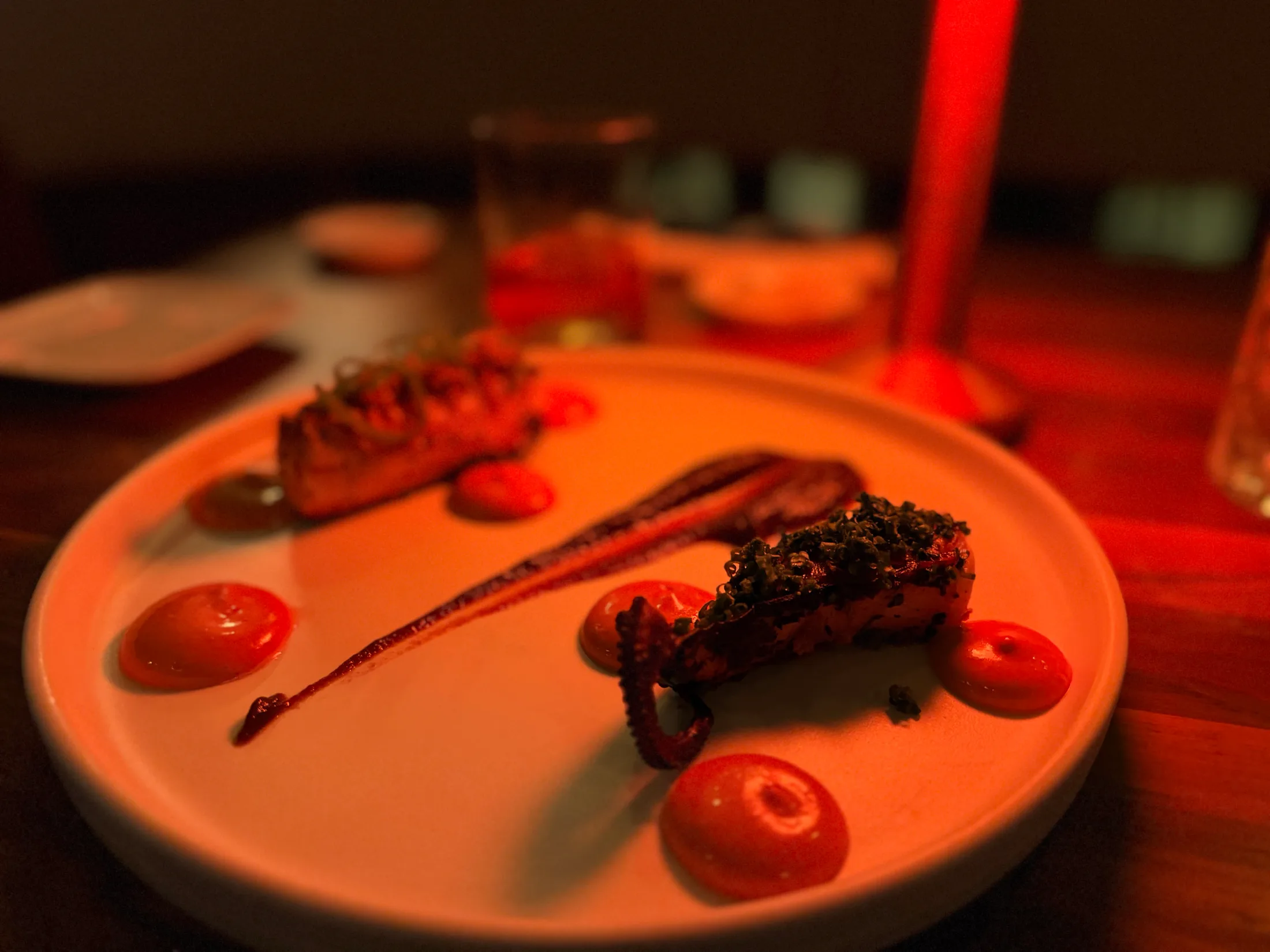 Composed octopus course plated on a white round plate with vivid red sauce dots, set against deep red ambient lighting