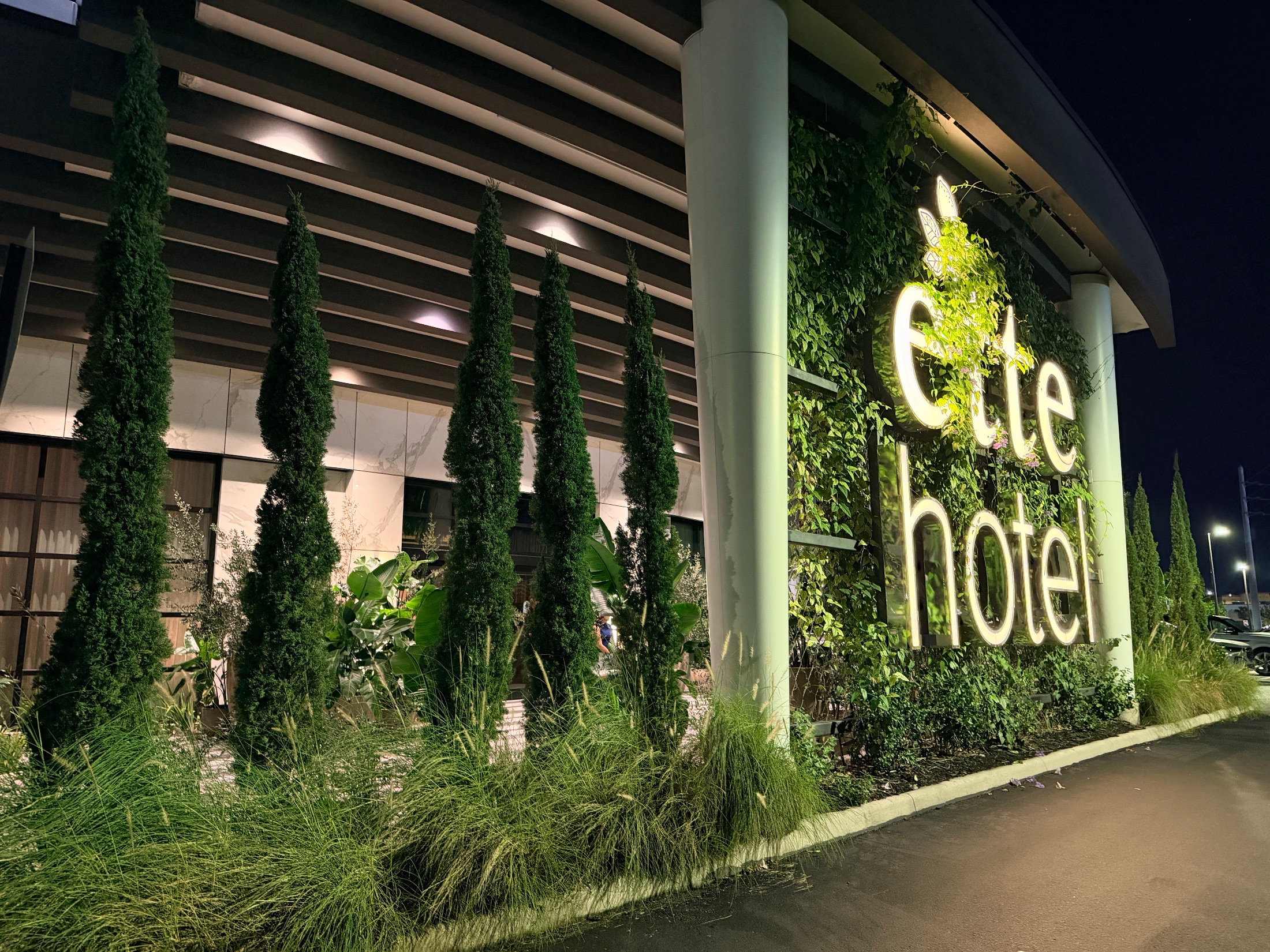 The illuminated ette hotel exterior at night, with a living green wall and tall topiaries lining the facade