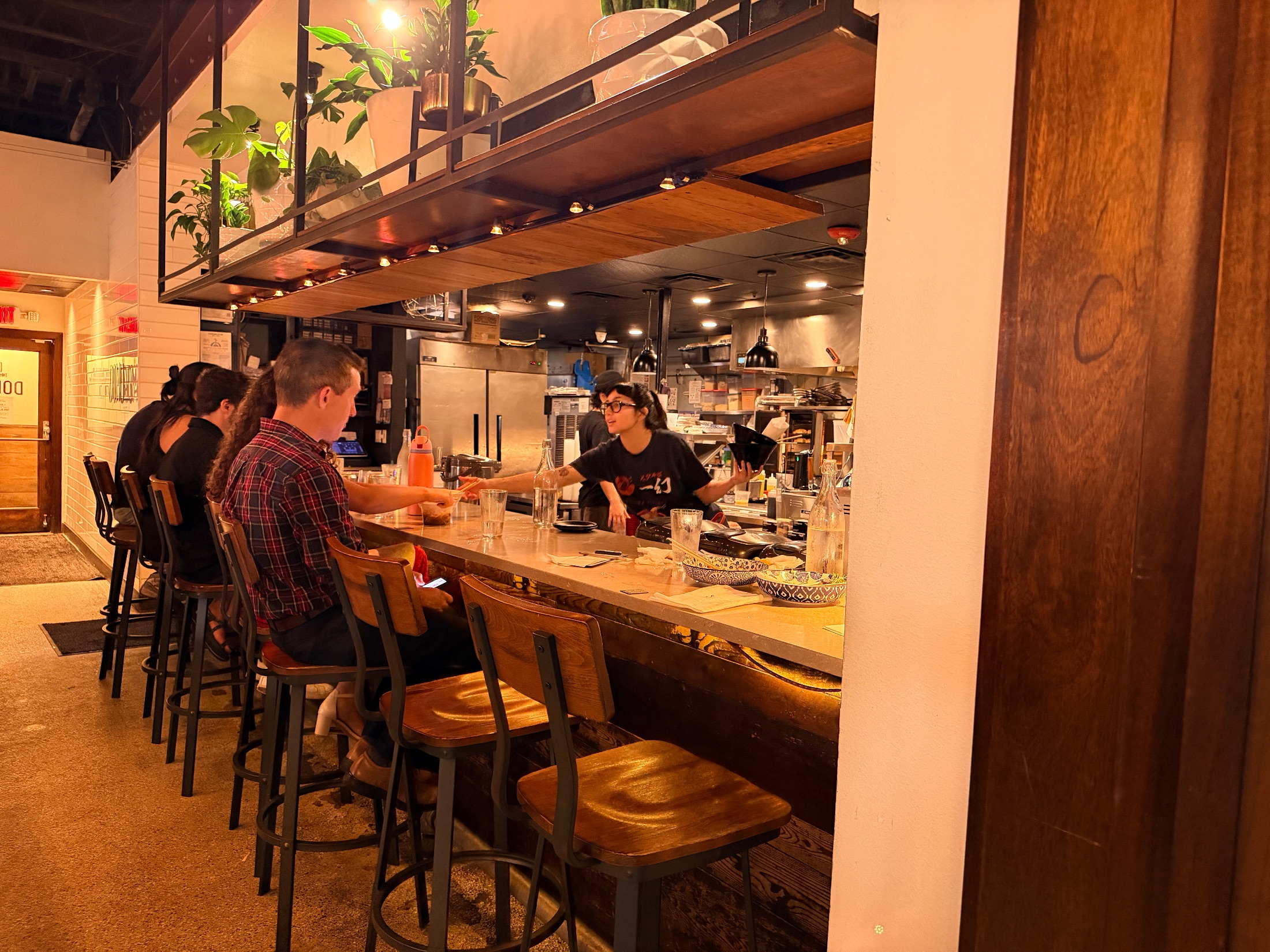 Wide shot of DOMU's bar area with Edison bulb ceiling installation, backlit whisky wall, and On Tap chalkboard in the background